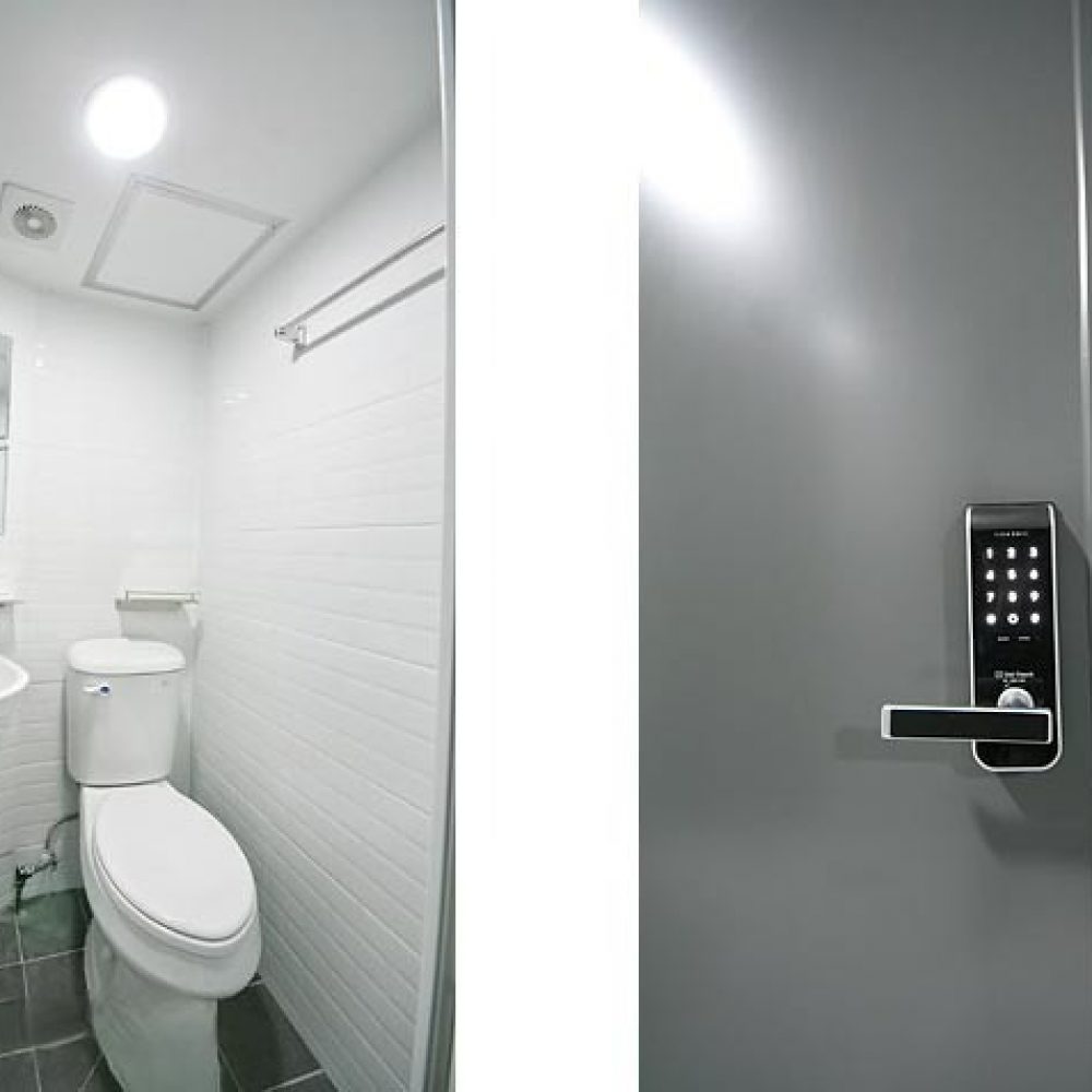 画像は、左側に白いタイルの浴室が映っています。洗面台とトイレがあり、鏡も設置されています。右側にはダークグレーのドアがあり、電子ロックのハンドルが取り付けられています。ドアの隣には石造りの壁が見え、全体的にモダンで清潔感のある印象を与えています。