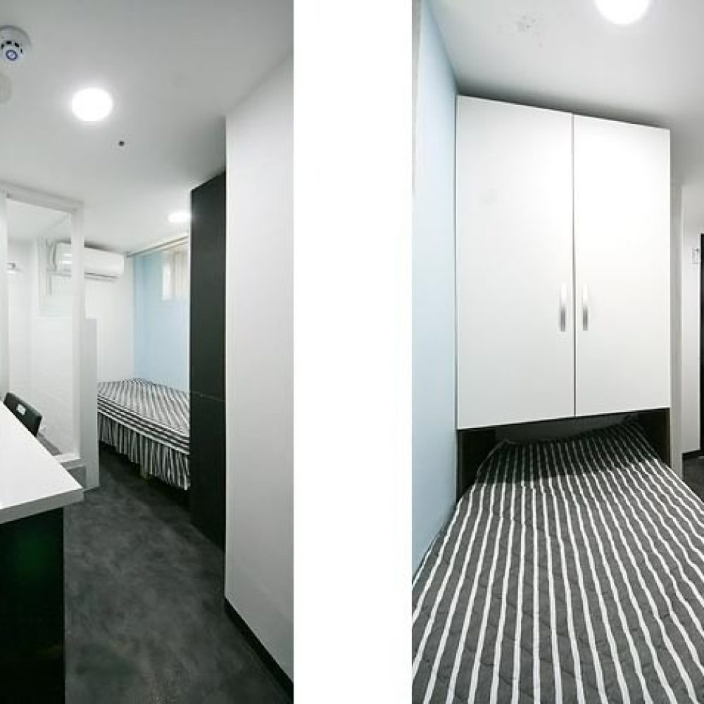部屋の内部が映った画像です。左側には白いカウンターといくつかの文房具が置かれたデスクがあります。奥にはストライプ模様のシーツがかけられたシングルベッドがあり、その向かい側には透明なガラスのシャワールームがあります。右側には収納キャビネットがあり、全体的にモダンで清潔感のあるインテリアが特徴です。