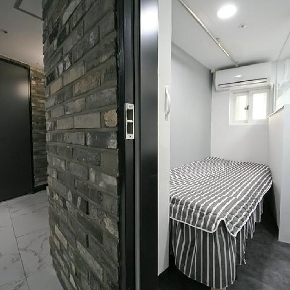 部屋の内部が映った画像です。左側にはレンガの壁があり、その先にダークカラーのドア（312と表示）が見えます。右側にはストライプ模様のシーツがかけられたシングルベッドがあり、さらに奥には透明なガラスのシャワールームとトイレがあります。全体的にモダンで清潔感のあるデザインです。