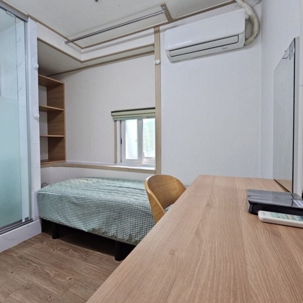 画像には、畳んだベッド、木製の机、エアコンなどが写っています。部屋は落ち着いた雰囲気で、整理整頓されたきれいな部屋に見えます。