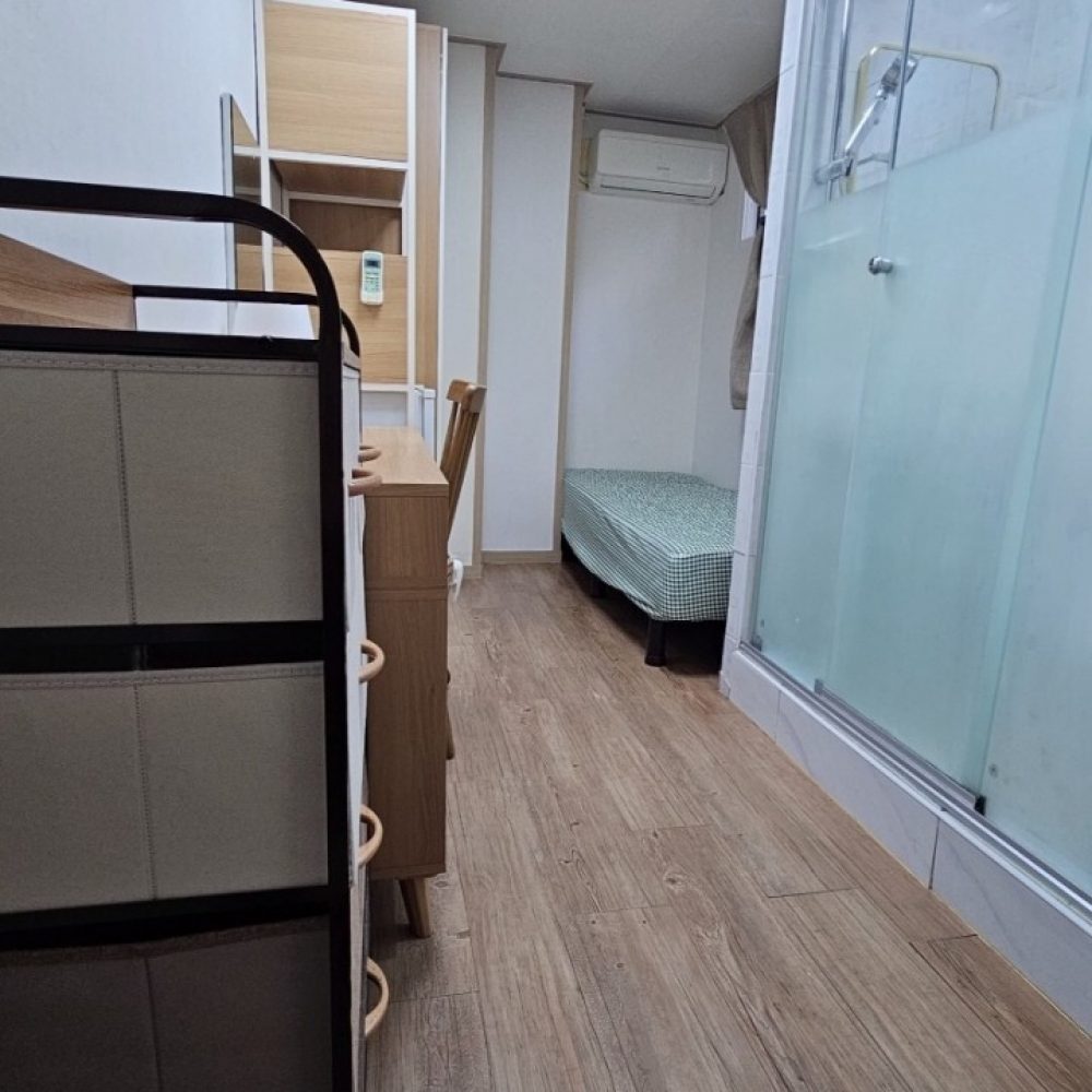 木製の床、棚、ベッドが見えるコンパクトな部屋の画像です。室内は明るく清潔感があり、収納スペースも確保されているようです。天井には照明が設置されており、快適な空間だと感じられます。