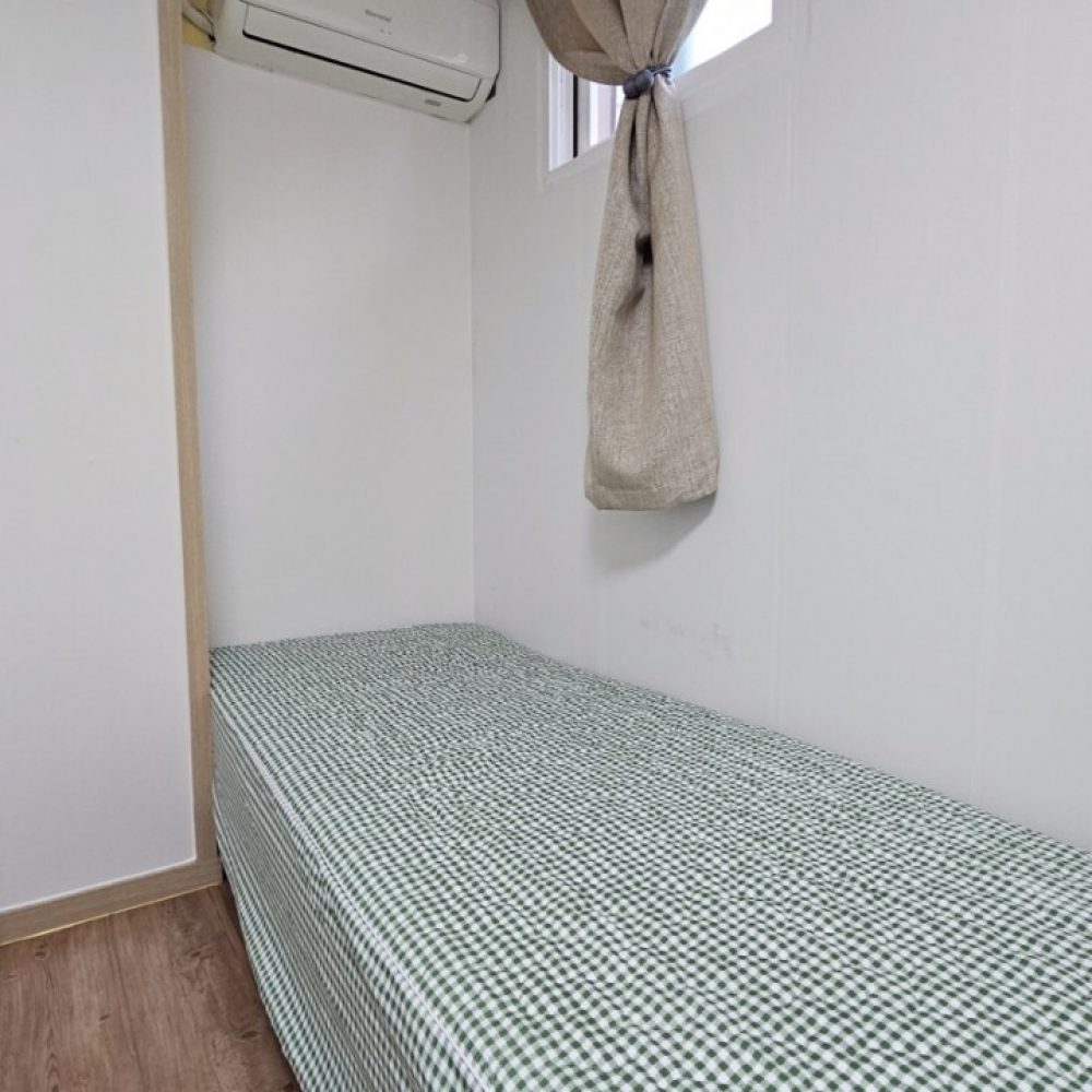 部屋の中の様子を示します。部屋には白い壁、木製の床、エアコン、そして窓の横に布の飾りがあります。部屋の中央には緑と白の格子模様のベッドリネンが敷かれた簡素なベッドが置かれています。全体として、シンプルながらも落ち着きのある雰囲気の部屋だと言えます。