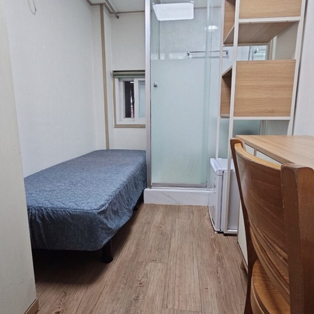 画像は小さな部屋の内部を写したものです。部屋には木目調の床、整理棚、ベッド、エアコン、冷蔵庫などが見られます。部屋は清潔で整理されており、居住者の生活スペースとして機能していると思われます。