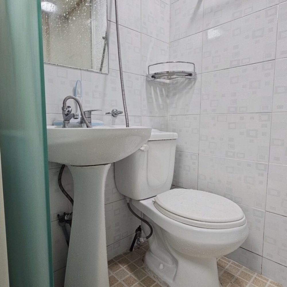浴室のイメージです。シャワーヘッドと洗面台、トイレがあり、タイルの壁と床で構成されています。トイレには蓋がついており、白を基調とした清潔感のある空間です。