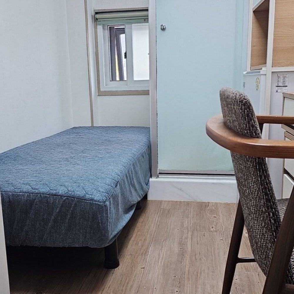画像は小さな居住空間を写したものです。ベッドや机、椅子などの家具が配置されており、シンプルで清潔な印象の部屋です。窓やシャワーカーテンなどの設備もあり、居住するのに適した空間のようです。