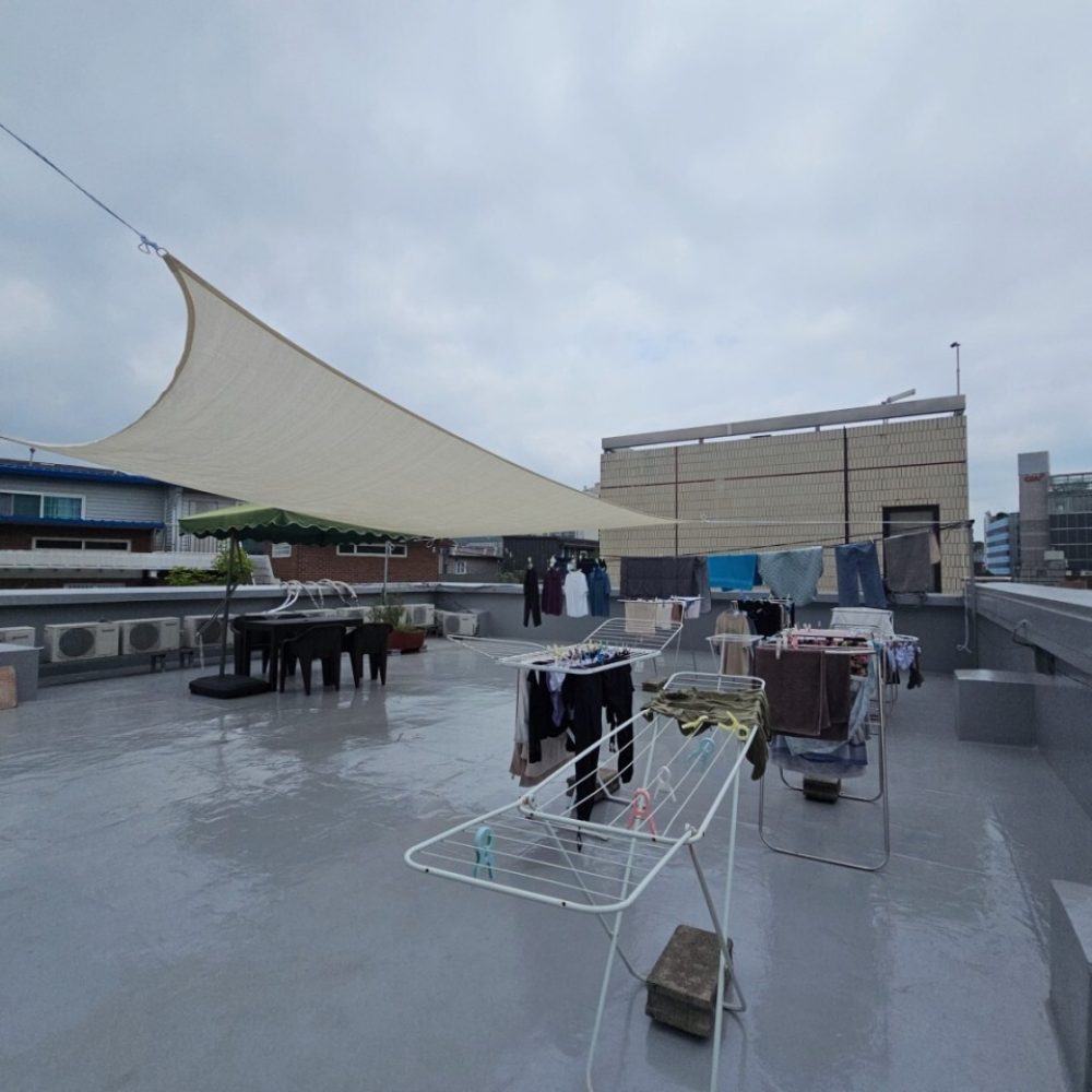 画像は屋外の建物の屋根の下にある洗濯物干し場の様子を捉えたものです。屋根は大きなサンシェードのようになっており、その下にはいくつかの洗濯物が干されています。建物の周りには、都市の高層ビルなども見えています。全体的に曇りの日の景色を表しています。