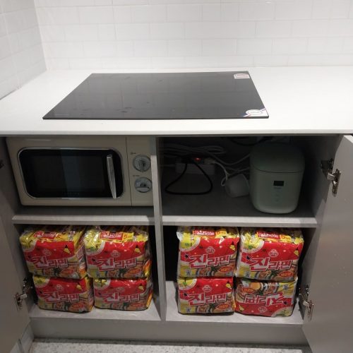 キャビネットの中には電子レンジやお米炊飯器といった調理家電、そして赤色の包装のお菓子類が整理されています。全体的に清潔で整理された印象の台所空間が写された画像です。