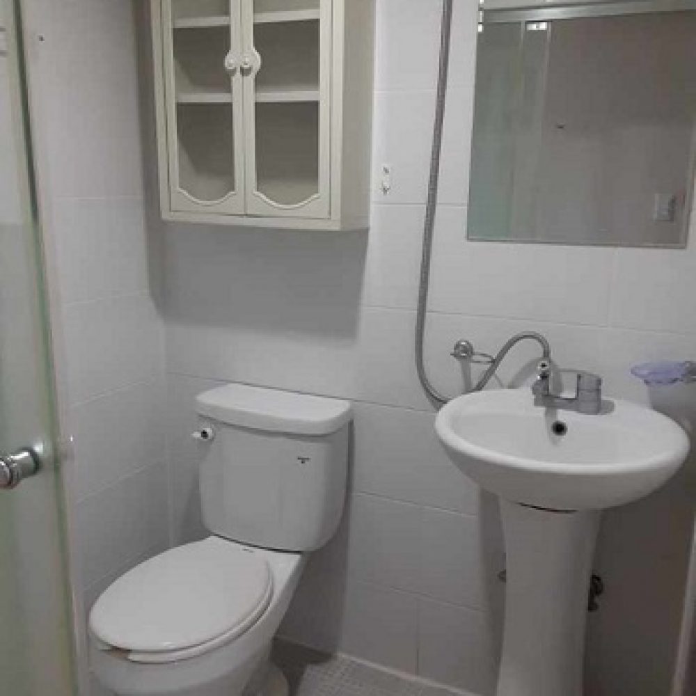 浴室の内部。白いタイルの壁と床が特徴で、トイレ、洗面台、鏡が設置されている。トイレは壁に沿って配置され、洗面台は手洗い用の蛇口が付いている。上部には収納キャビネットがあり、全体的に清潔感のある空間が広がっている。
