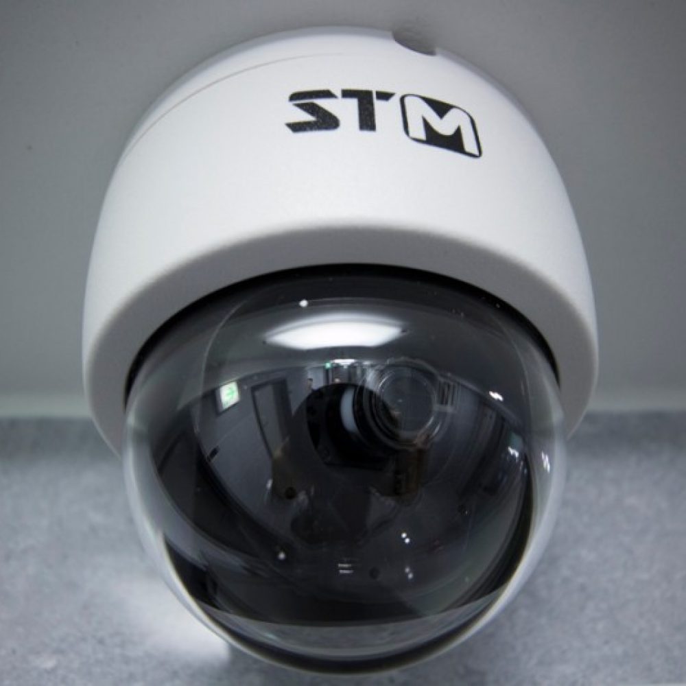 カメラのドーム型セキュリティカメラ。上部に「STM」というロゴがあり、透明なレンズが特徴的です。背景はグレーのタイルのように見えます。