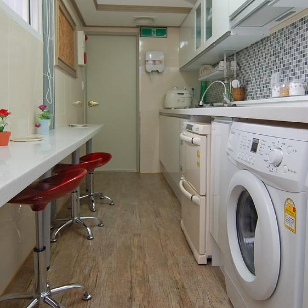 キッチンと洗濯機が並んだ部屋の内部。左側にはコンピュータと赤い椅子が並び、机の上には小さな花が飾られている。右側には2台の洗濯機と料理家電が配置されています。明るい色調のインテリアで、清潔感のある空間。