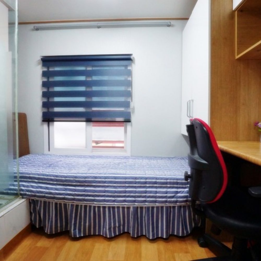 部屋の内部が映った画像です。ストライプ模様の布団が掛けられたベッドがあり、背の高いデスクには黒い椅子が置かれています。窓には青いブラインドがあり、ガラスの壁で仕切られたエリアが見えます。また、デスクの上には小さな花が飾られた容器もあります。全体的に明るく清潔感のある雰囲気です。