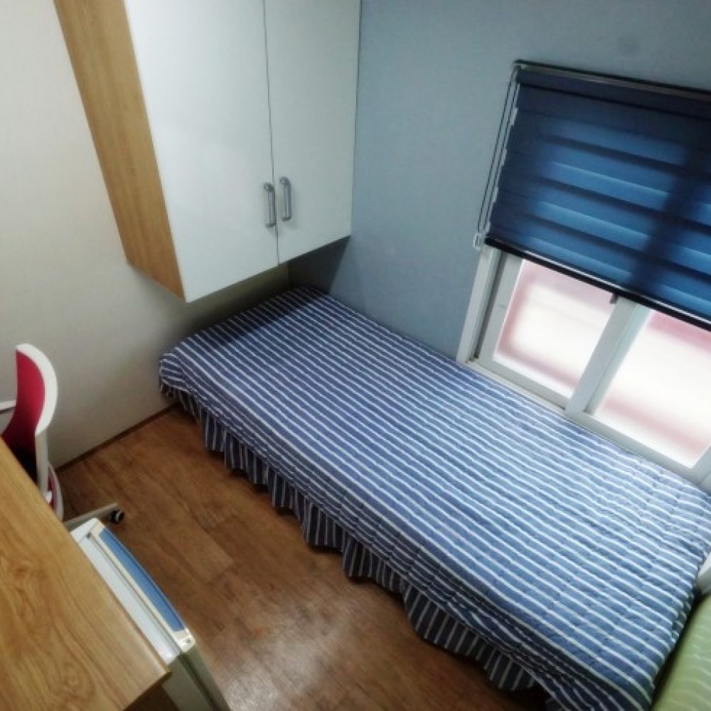 部屋の俯瞰図が映った画像です。ストライプ模様の布団が掛けられたベッドがあり、その横には木製のデスクと椅子があります。壁には収納キャビネットがあり、窓にはブルーのブラインドがあります。床は木目調で、全体的に明るい雰囲気の部屋です。