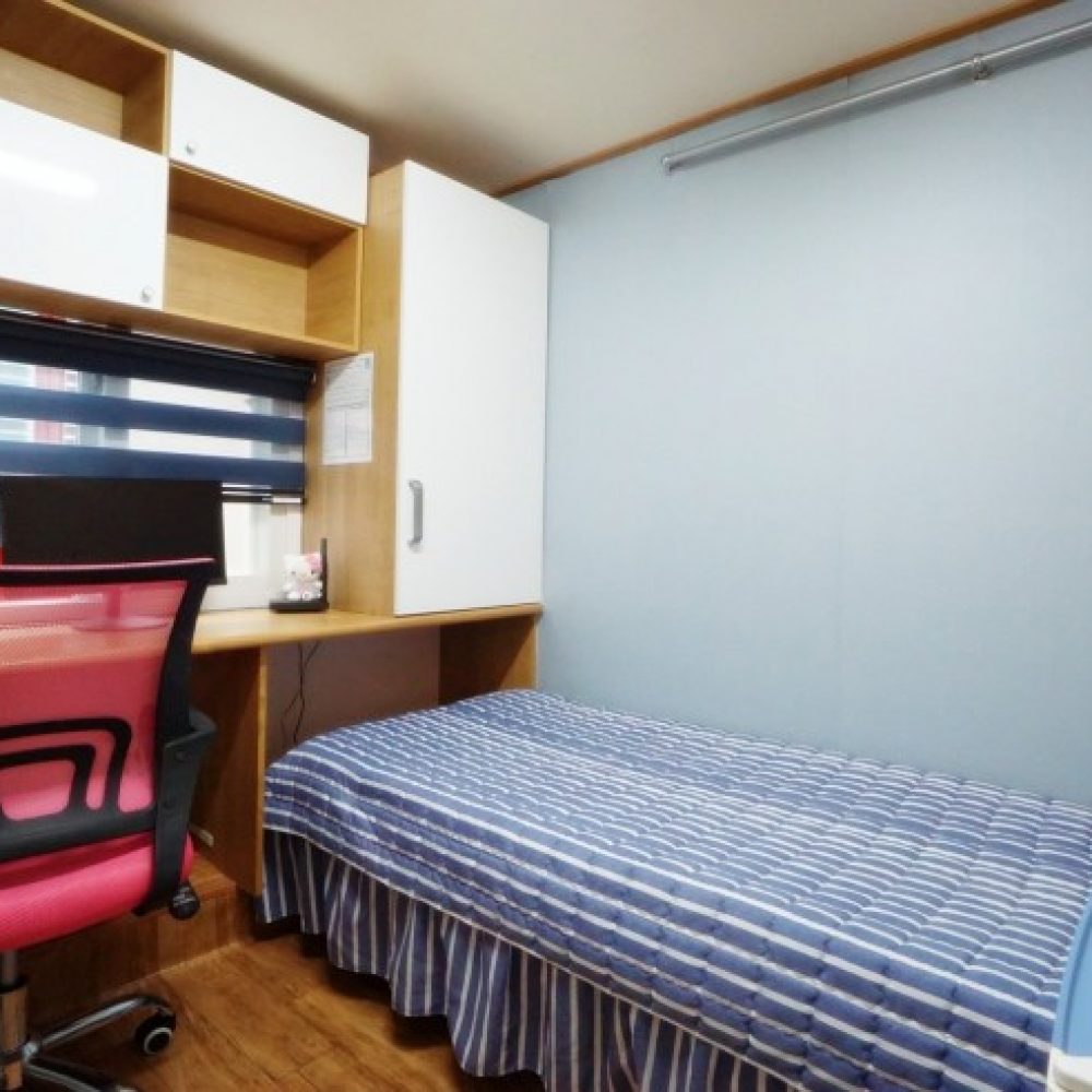 部屋の内部が映った画像です。シンプルなデザインのベッドがあり、ストライプ模様の布団が掛けられています。ベッドの隣には木製のデスクがあり、その上には黒い椅子が置かれています。窓にはブラインドがあり、壁は淡い青色です。壁には収納キャビネットもあります。全体的に落ち着いた雰囲気のある空間です。