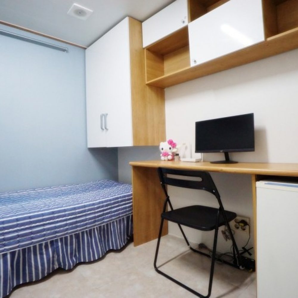 部屋の内部が映った画像です。ストライプ模様の布団が掛けられたベッドと、木製のデスクがあります。デスクの上には黒いモニターが置かれ、椅子は折りたたみ式です。また、壁には収納キャビネットがあり、小型の冷蔵庫も見えます。全体的にシンプルで明るい雰囲気の部屋です。
