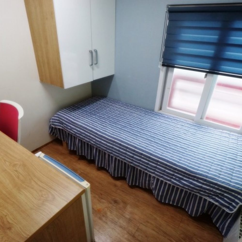 部屋の内部が映った画像です。ストライプ模様の布団が掛けられたベッドがあり、その横には木製のデスクとピンクの椅子があります。壁には収納キャビネットが取り付けられ、窓には青いブラインドがあります。床は木目調で、全体的に明るく清潔感のある雰囲気です。