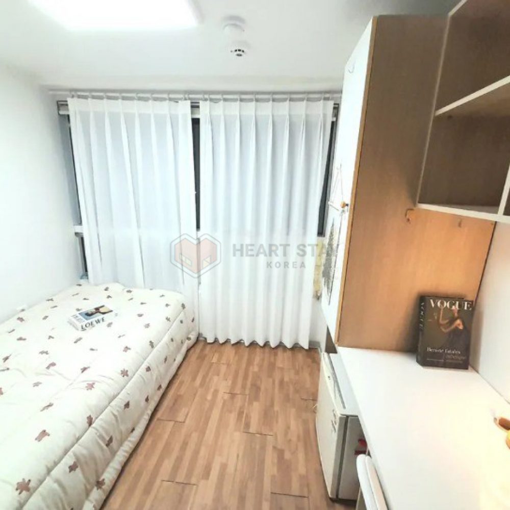 この画像は、落ち着いた雰囲気の寝室を示しています。部屋の中には白いカーテンが掛かっており、日光がやわらかく差し込んでいます。中央には、花柄のシーツで覆われたシングルベッドがあり、その上には雑誌が置かれています。右側には、収納キャビネットとデスクエリアがあり、デスク上には「VOGUE」と書かれた本と小さなランプがあります。全体的にシンプルで明るい印象の部屋です。